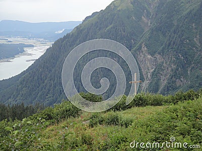 Cross on Mountain Range with Forests and Stream. Lush temperate rainforest in alaska with clouds and sun Stock Photo