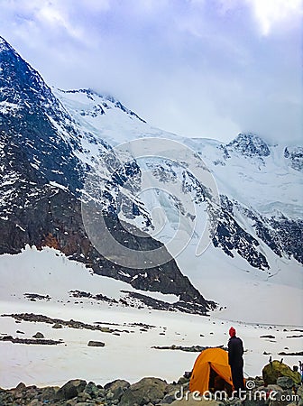 The mountaineer stands at the tent at the foot of a snow-covered mountain. Concept of travel and extreme relaxation. Stock Photo