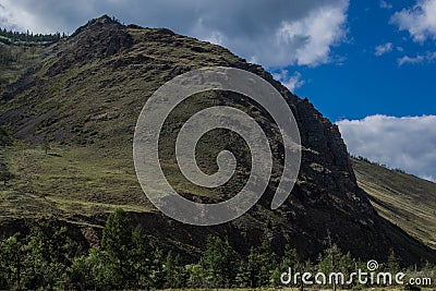 Mountains on Baikal, Sarma. Large tall green grassy round rock. Trees and bushes in the foreground in dry steppe. Cloudy blue sky Stock Photo