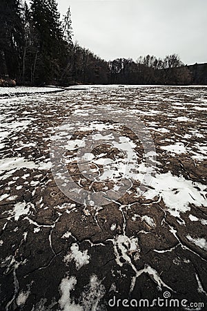 Muddy and snow covered cracked ground earth with forest stream in winter. Wide angle photo of snowy and dirty river in forest - Stock Photo