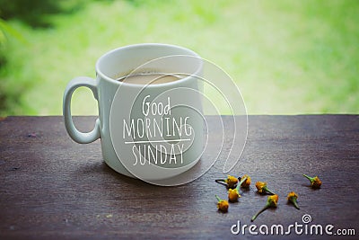 Mug of coffee. Morning white coffee with greeting text on it - Good Morning Sunday, and yellow little flowers arrangement. Stock Photo