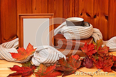 Mug of coffee wrapped in gray woolen scarf, empty wooden frame, autumn leaves and stack of books on wooden table Stock Photo