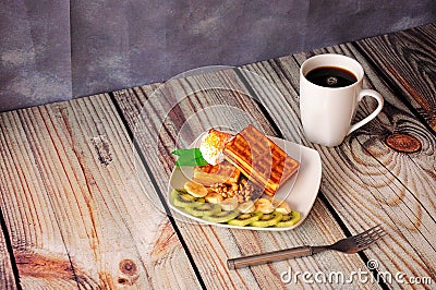 A mug of hot black coffee and a plate with waffles with cream, kiwi and banana slices on a wooden table Stock Photo