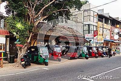 Multi-colored tuk-tuks stand in a row, near the road next to buildings Editorial Stock Photo