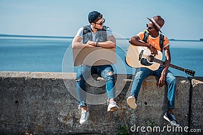 multicultural young men with cardboard and guitar resting on parapet while hitchhiking Stock Photo