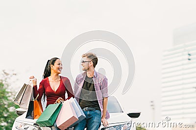 Multiethnic couple with shopping bags, smiling and sitting on white car. Love, casual lifestyle, or shopaholic concept Stock Photo