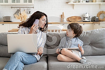Busy businesswoman mother work from home talk to kid with laptop on knees calling on phone on couch Stock Photo