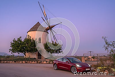 Murcia, Spain - October 26, 2019: A red Tesla Model 3 is parked in front of a windmill surrounded by trees. Its headlights are on Editorial Stock Photo