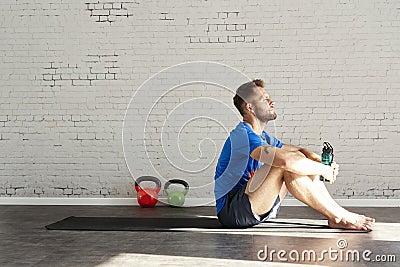 Muscular athlete sitting in sunny sport club, relaxed after workout, bottle of water in hands. Space for text layout on brick wall Stock Photo