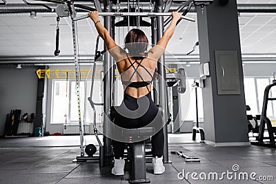 Muscular athletic female bodybuilder in black suit pulled on sports simulator in gym. Back muscles are very tense Stock Photo