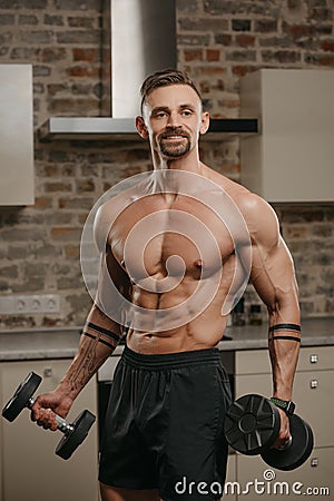 A muscular man with a beard is posing with dumbbells in his apartment Stock Photo