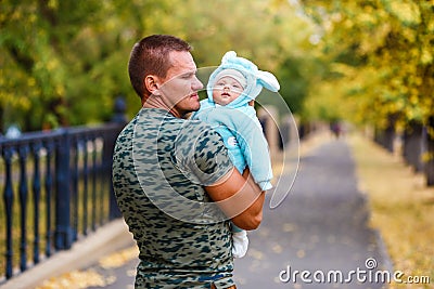 A muscular man holds the baby in warm jumpsuit in the fall on a walk. yellow leaves Stock Photo