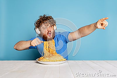 Excited young hairy red-bearded man tasting large portion of noodles, pasta isolated on blue studio background. Stock Photo