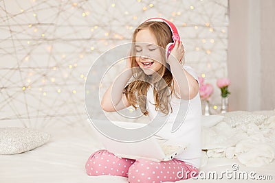 Music and technology. Portrait of young girl in pajamas with white laptop and wireless pink headphones, sing song on Stock Photo