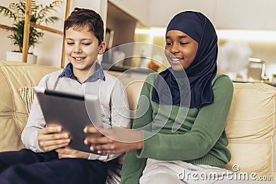 Muslim girl and her caucasina friend sitting on sofa at home using digital tablet Stock Photo