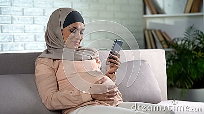 Muslim pregnant female watching video on smartphone home, expecting childbirth Stock Photo