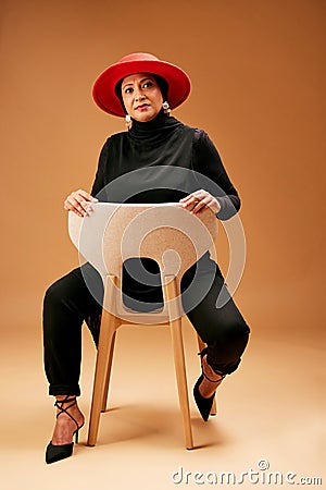 Muslim woman, chair and fashion, beauty and hat with trendy outfit, focus attitude and studio background. Mature Stock Photo