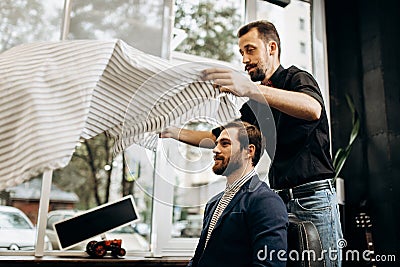 Mustachioed barber dressed in a black shirt puts a cape on the stylish man sitting in the armchair in a barbershop Stock Photo