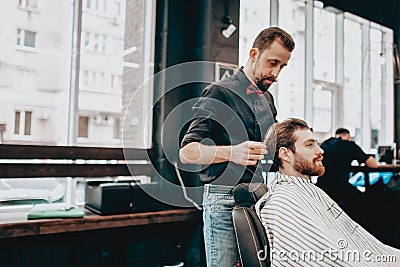 Mustachioed barber dressed in a black shirt with a red bow tie makes a stylish hairstyle to young man in a barbershop Stock Photo