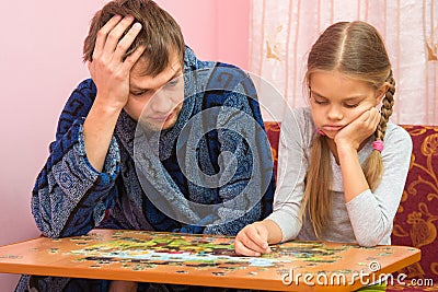 My daughter collects picture from puzzles, tired dad sitting next to her and tells Stock Photo
