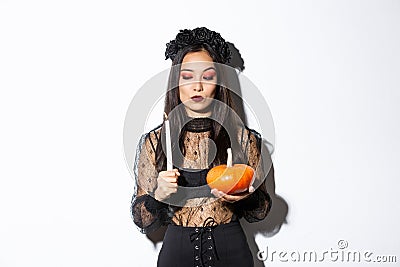 Myserious asian wicked witch in gothic dress, looking at lit candle, holding pumpkin, standing over white background Stock Photo