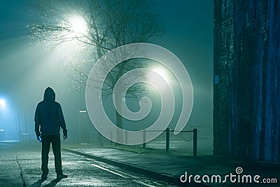 A mysterious hooded figure holding a phone, standing in a city street. On a moody foggy winters night Stock Photo