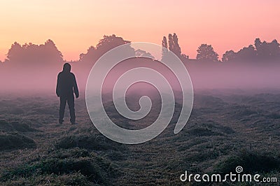 A mysterious hooded, figure standing in a field on a beautiful early misty morning, looking at the sunrise Stock Photo