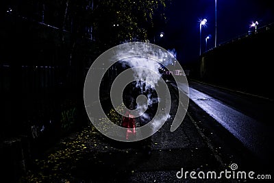 Mysterious man exhaling vaping smoke which hides its face while walking on the street during nighttime Stock Photo