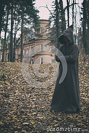 Mysterious monk, wizard or adept of secret society in hooded robe stands in forest against the background of medieval castle or Stock Photo