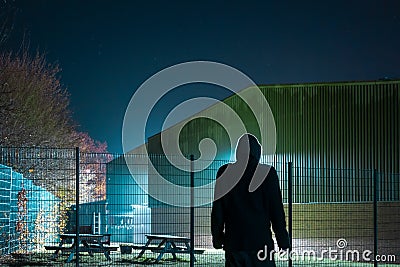 A mysterious, scary hooded figure. Silhouetted by back light. Looking at a warehouse on a winters night. Stock Photo