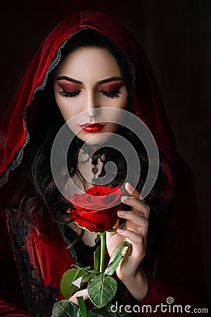 Mystical beautiful woman in a gothic costume of a medieval vampire in a hood. An adult girl holds a rose flower in her Stock Photo