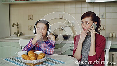 Mãe nova e filha bonito que comem o café da manhã na manhã na cozinha em casa Música de escuta da menina com video estoque Mãe nova e filha bonito que comem o café da manhã na manhã na cozinha em casa Música de escuta da menina com video estoque