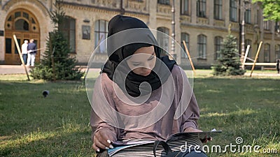 Młoda muzułmańska dziewczyna w hijab siedzi na gazonie i czytelniczy magazyn, builging na tle, religijny pojęcie, relaksuje zbiory wideo Młoda muzułmańska dziewczyna w hijab siedzi na gazonie i czytelniczy magazyn, builging na tle, religijny pojęcie, relaksuje zbiory wideo