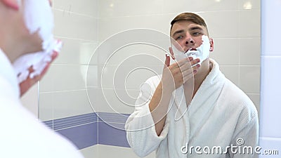 Młody mężczyzna stosuje pianę golenia przed lustrem Mężczyzna w białym płaszczu z pianą na twarzy Wyświetl przez zbiory wideo Młody mężczyzna stosuje pianę golenia przed lustrem Mężczyzna w białym płaszczu z pianą na twarzy Wyświetl przez zbiory wideo
