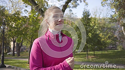 Nahaufnahme eines lächelnden kaukasischen Mädchens in rosa Sportbekleidung und Kopfhörer im Herbstpark Frauenläufer stock footage Nahaufnahme eines lächelnden kaukasischen Mädchens in rosa Sportbekleidung und Kopfhörer im Herbstpark Frauenläufer stock footage