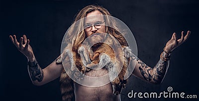 Naked tattoed redhead hipster male with long luxuriant hair and full beard posing with the fox skins on his shoulders in Stock Photo