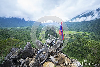 NAM XAY VIEWPOINT, VANG VIENG, LAOS - JULY 25, 2019: Ancient motorcycle parked and Lao flag on the Nam Xay Viewpoint, During the Editorial Stock Photo