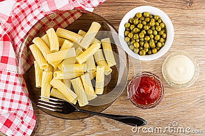 Napkin, bowls with green peas, mayonnaise, ketchup, brown plate with cooked pasta, fork on table. Top view Stock Photo