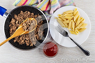 Napkin, spoon in frying pan with fried mince, white plate with pasta, fork, bowl with ketchup on table. Top view Stock Photo