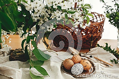 Natural Easter eggs painted with wax on vintage plate on background of wicker basket, white spring flowers and greenery on wooden Stock Photo