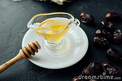 Natural honey in transparent saucepan with spout and wooden honey stick on saucer on dark table with dried dates scattered around Stock Photo