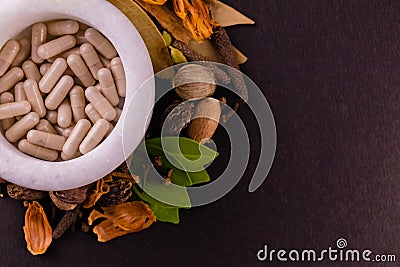 Natural medicine concept. Top view of ayurvedic capsules in a mortar and different spices, green leaves on black background with Stock Photo