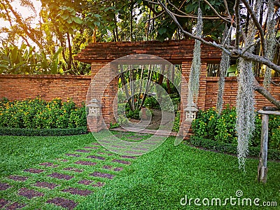 Natural orange clay brick arched wall entrance in a tropical garden with pattern of brown laterite walkway on green grass lawn Stock Photo