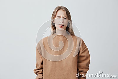 Naughty beautiful girl with blonde long hair in beige sweater misbehaving, sticking out tongue at camera as a sign of Stock Photo