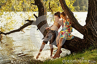 Naughty boy and girl sitting on a branch over water, laughing, having fun talking Stock Photo