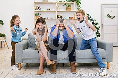 Naughty Kids Shouting Loudly Standing Near Tired Parents At Home Stock Photo