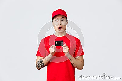 Nervous asian delivery guy in red t-shirt and cap uniform, holding credit card, staring camera and gasping. Young Editorial Stock Photo