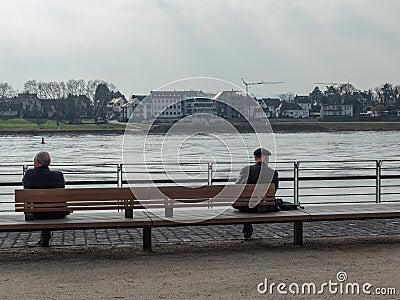 Neuwied, Germany - March 23, 2021: two men seen from behind, sitting with distance on a bench at the quayside of the Rhine Editorial Stock Photo