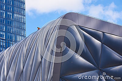 A man in an orange shirt works on the roof of The Shed. He is dwarfed by the huge size of the building and the glass skyscraper Editorial Stock Photo