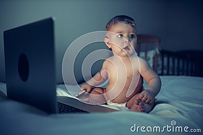 A newborn baby uses a computer while sitting on a sofa at night. The child is dressed in a diaper on his body Stock Photo